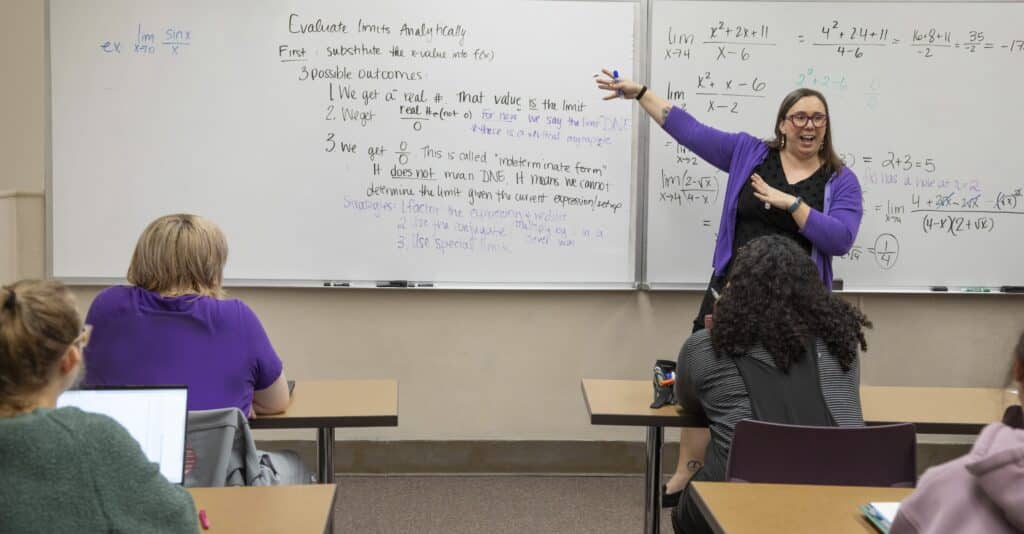 A female professor stands in front of a classroom pointing to a whiteboard filled with mathematics notes and formulas. Students are shown from behind listening intently.