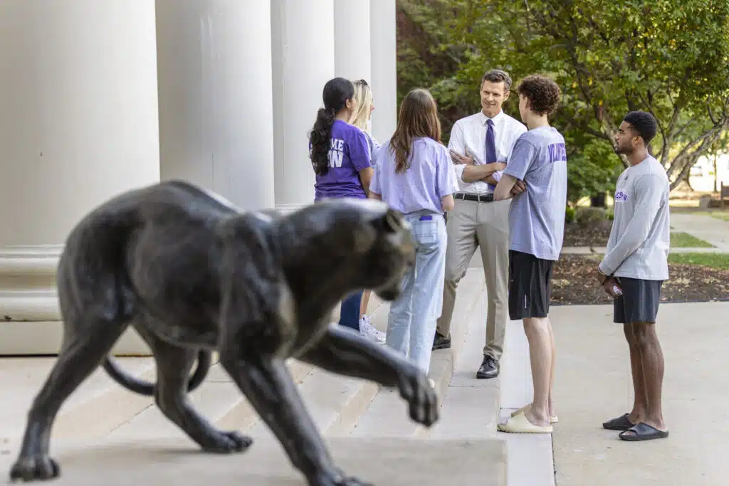 University President Dr. Cousins speaking with a group of students in the background, with the panther statue in the foreground outside on campus
