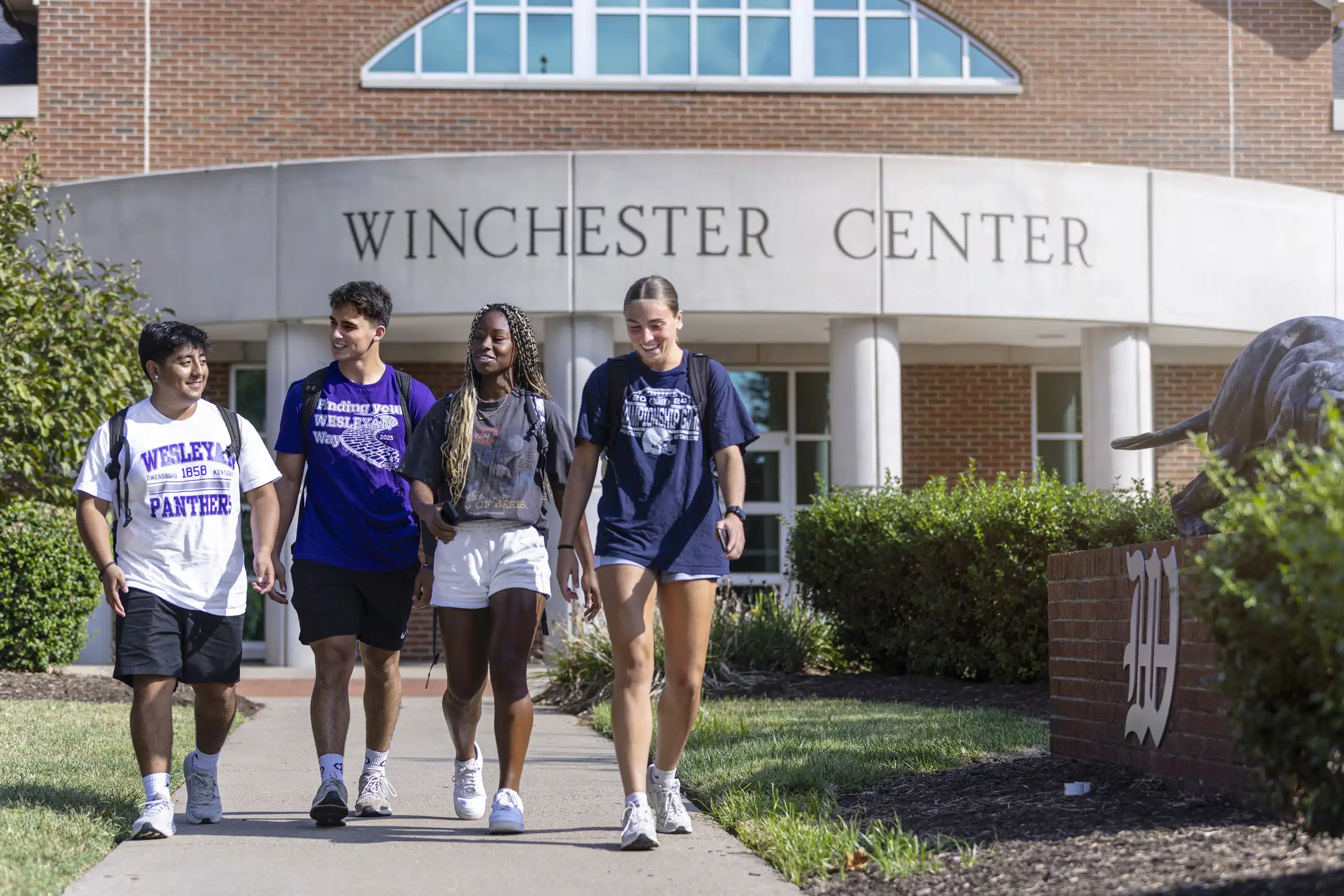 A group of four KWC students walking on the sidewalk outside the Winchester Center on campus