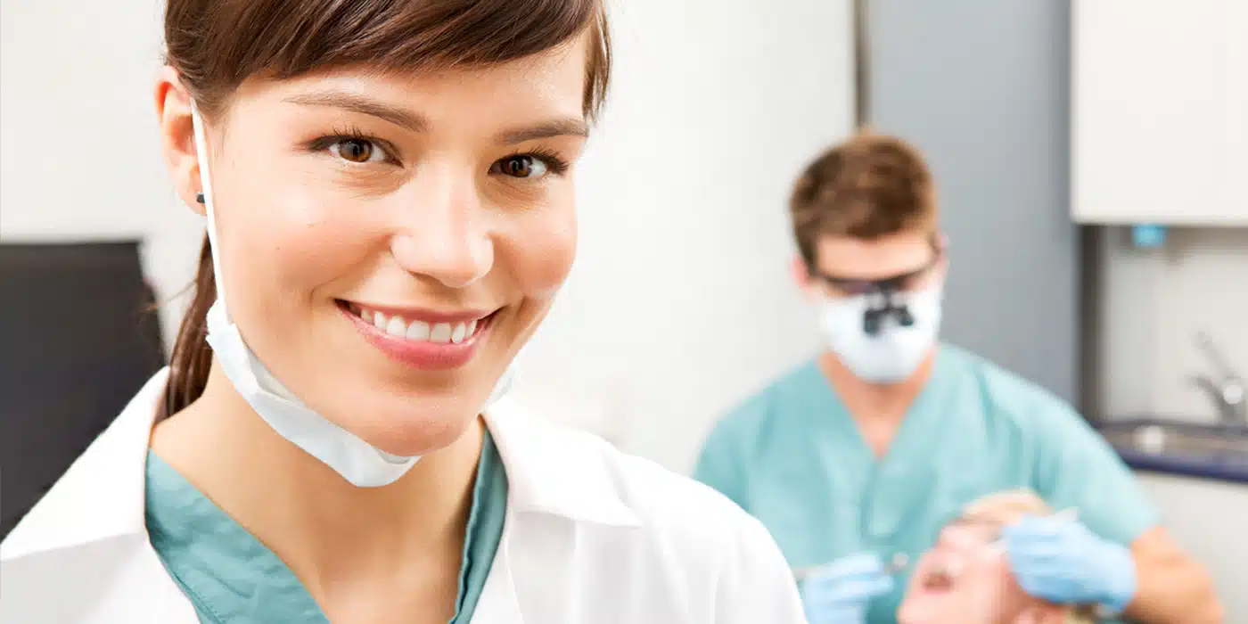 Smiling dental assistant with a dentist treating a patient in the background